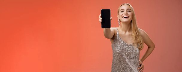 Stolz freudig charmant fröhlich blond europäisch Frau in stilvollem silber glänzenden Kleid halten Hand Taille selbstbewusst ausstrecken Arm zeigt Smartphone-Display präsentieren awesome neues App-Gerät, roter Hintergrund. - Foto, Bild