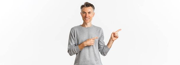 Image of handsome middle-aged man with grey short hairstyle, smiling pleased as pointing fingers left, showing logo on copy space, standing over white background. - Photo, Image