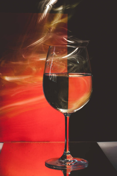 Csendélet egy pohár vízzel és fényvonalakkal körülvett színes vörös és fekete háttér, amely létrehoz egy illúzió. - Fotó, kép