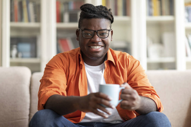 Sonriente guapo gordito negro en traje casual sentado en el sofá con taza de té de hierbas, chico afroamericano positivo bebiendo café en acogedora sala de estar en casa, mirando a la cámara, espacio de copia - Foto, Imagen