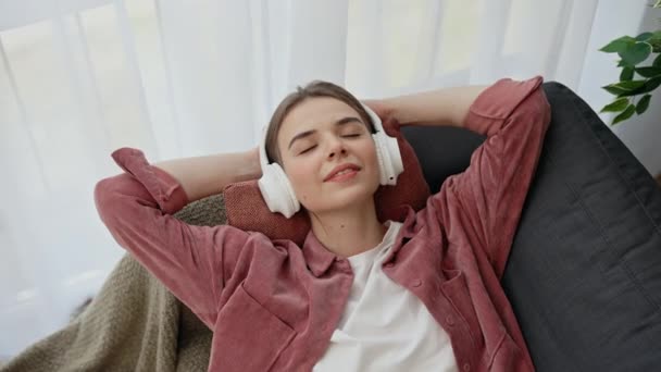 Relajada mujer caucásica está tumbada en el sofá en casa y disfrutando de escuchar música con los auriculares. Concepto de personas y tecnología - Imágenes, Vídeo
