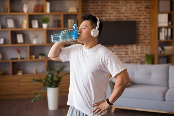 Sportovní dospělý korejský muž se sluchátky pitnou vodou po tréninku doma, hydratovaný během domácího sportovního tréninku. Zdravý životní styl, wellness koncept - Fotografie, Obrázek
