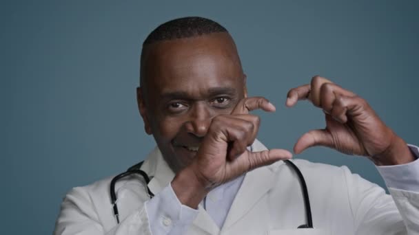 Взрослый улыбающийся мужчина врач профессиональный кардиолог в медицинском халате показывая руки в форме сердца жест любви глядя на камеру медицинского страхования реклама кардиологической службы благотворительной поддержки - Кадры, видео