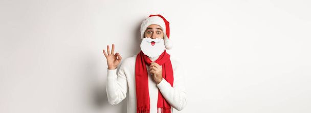 Χριστουγεννιάτικο πάρτι και εορταστική ιδέα. Χαρούμενο ανδρικό μοντέλο σε καπέλο Αϊ Βασίλη και λευκή μάσκα γενειάδας, που δείχνει ok χειρονομία, στέκεται πάνω από το λευκό φόντο. - Φωτογραφία, εικόνα