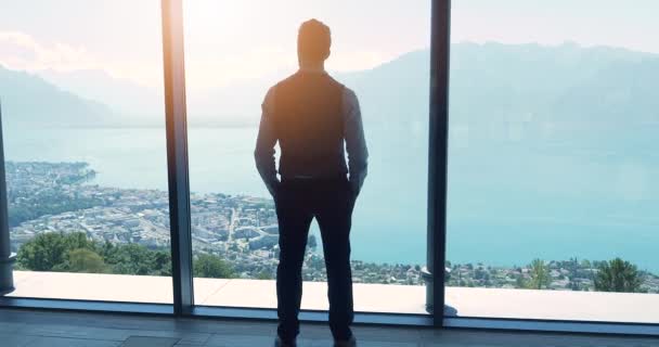 Молодой бизнесмен в современном офисе смотрит в окно с захватывающим видом на озеро и горы  - Кадры, видео