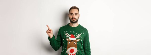 Χειμερινές διακοπές και Χριστούγεννα. Όμορφος γενειοφόρος άνδρας σε πράσινο πουλόβερ δείχνει το δάχτυλο δεξιά, δείχνει promo, στέκεται πάνω από το λευκό φόντο. - Φωτογραφία, εικόνα