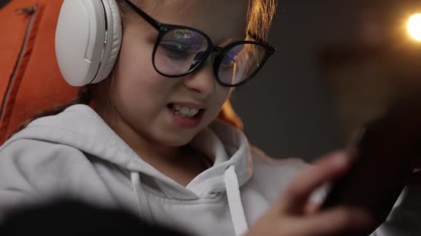 Возбужденная девочка-подросток играет в игры дома на цифровых планшетных технологиях устройства, сидя на диване. Ребенок в наушниках и очках держит в руках планшет для серфинга в Интернете.. - Кадры, видео