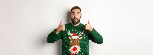 クリスマス、休日やお祝い。白い背景の上に立って、偉大な何かのような承認で親指を示す、緑のセーターの若い男を驚かせた。. - 写真・画像