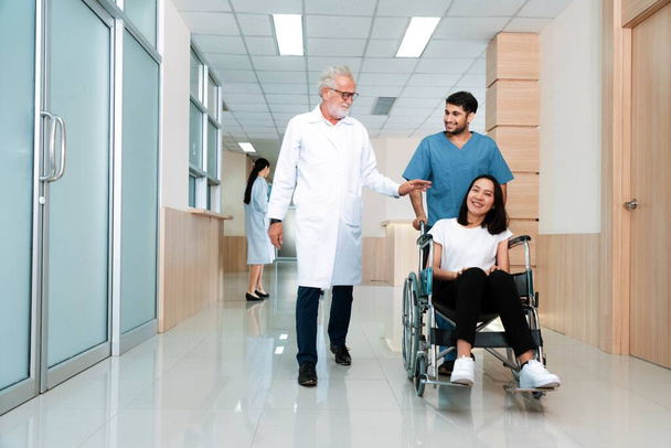 Lekarz i pielęgniarz przewożą pacjentkę na wózku inwalidzkim sterylnym szpitalnym korytarzem. Opieka zdrowotna i opieka pielęgniarska nad niepełnosprawnym pacjentem w koncepcji szpitala. - Zdjęcie, obraz