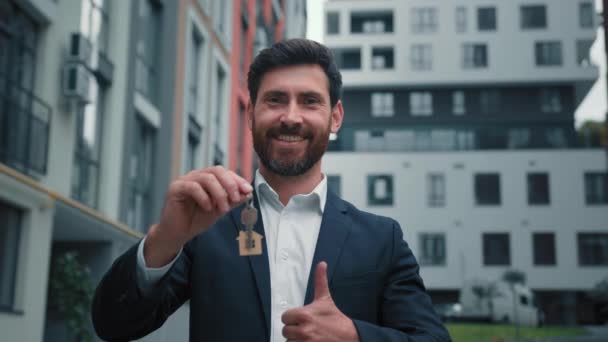 Szczęśliwy właściciel domu kaukaski mężczyzna nieruchomości agent nieruchomości przedsiębiorca zwycięzca trzymać klucze z nowego mieszkania sprzedać mieszkanie wygrać nieruchomości mieszkaniowej zrobić kciuk w górę gest polecam dom hipoteka oferta kredyt bankowy - Materiał filmowy, wideo