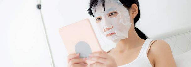Schöne junge asiatische Frau mit Laken-Gesichtsmaske sitzt vor dem Tablet-Computer auf dem Bett im Schlafzimmer, Schönheit Mädchen Anwendung Gesichtsmaske für Hautpflege von Falten zu Hause, Hautpflege, Gesundheit und Wohlbefinden. - Foto, Bild