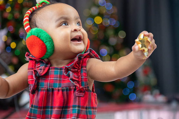 O bebê afro-americano está sorrindo alegremente enquanto brinca com a bugiganga do ornamento enquanto se veste com o vestido de Natal e o chapéu de Papai Noel com a árvore de Natal na parte de trás para a celebração da temporada - Foto, Imagem