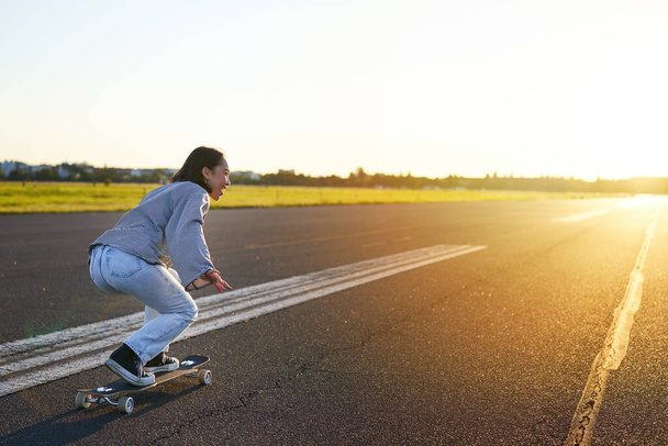 Zijaanzicht van mooi Aziatisch meisje op skateboard, rijdend op haar cruiser naar de zon op een lege weg. Gelukkig jong skater genieten van zonnige dag op haar skate. - Foto, afbeelding