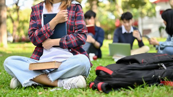 Csinos fiatal ázsiai egyetemista lány alkalmi ruhában, kezében a tabletjével, ül a füvön a gyönyörű egyetemi parkban. kivágott kép - Fotó, kép