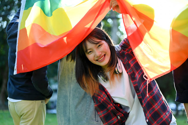 Schöne und glückliche junge asiatische Studentin, die mit ihren Freunden die LGBT-Regenbogenfahne hält. Einheit von Minderheit, Freiheit und Gleichheit, LGBTQ + Stolz. - Foto, Bild