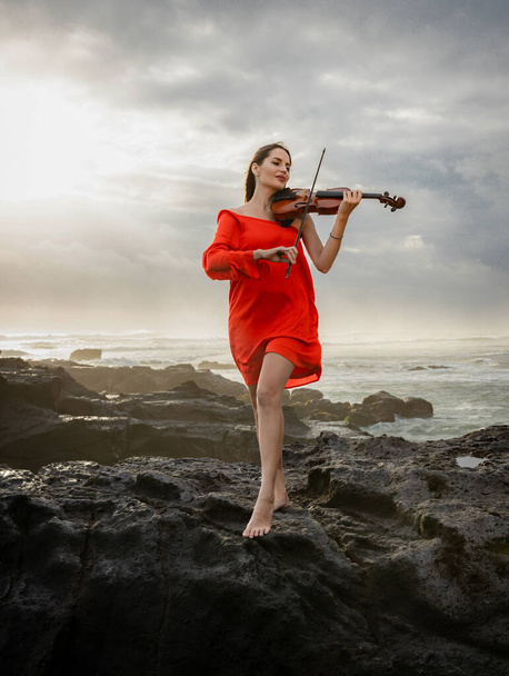 Attractive woman with violin on the beach. Music and art concept. Slim girl wearing short red dress and playing violin in nature. Sunset time. Cloudy sky. Bali, Indonesia - Photo, Image