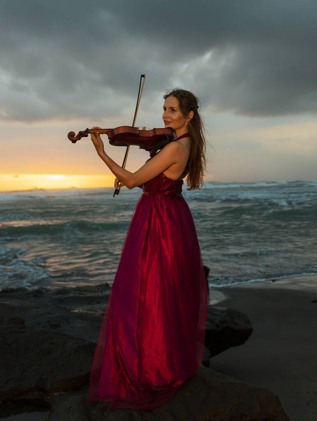 Charming Caucasian woman with violin on the beach. Music and art concept. Slim girl wearing long red dress and playing violin in nature. Sunset time. Cloudy sky. Bali, Indonesia - Photo, Image