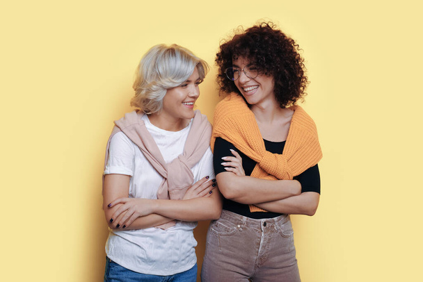 Portretfoto van twee jonge vrouwen die met gekruiste armen naar elkaar staan te kijken terwijl ze glimlachen op een geïsoleerde beige achtergrond. Goede vrienden en team - Foto, afbeelding