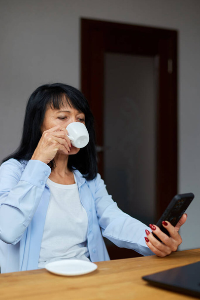 Iäkäs nainen liikenainen rentoutuu työpaikalla, käyttää matkapuhelinta ja juo kahvia tauolla virka-aikana. Naisten lepo työpaikalla - Valokuva, kuva
