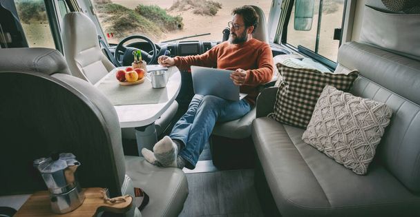 Un solo hombre sentado y relajante dentro de una furgoneta de autocaravana alternativa estilo de vida en casa fuera de la red utilizando un ordenador portátil y tomando un café en total libertad de relajación. Motor interior casa actividad de ocio de viajes - Foto, imagen