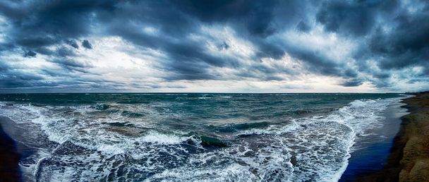 Δραματικός συννεφιασμένος ουρανός με σύννεφα νιμποστράτου και κακοκαιρία σε πανοραμική θέα στην παραλία με θυελλώδη κύματα που απειλούν να συντριβούν στην ακτή με πολύ θαλασσινό αφρό. - Φωτογραφία, εικόνα