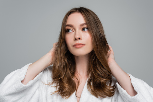 young woman in bathrobe touching shiny and healthy hair while looking away isolated on grey - Foto, imagen