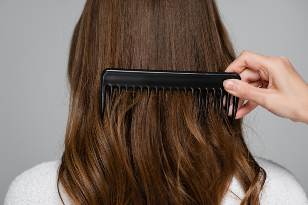 przycięty widok fryzjer szczotkowanie błyszczące i zdrowe włosy młodej kobiety z grzebieniem izolowane na szary - Zdjęcie, obraz