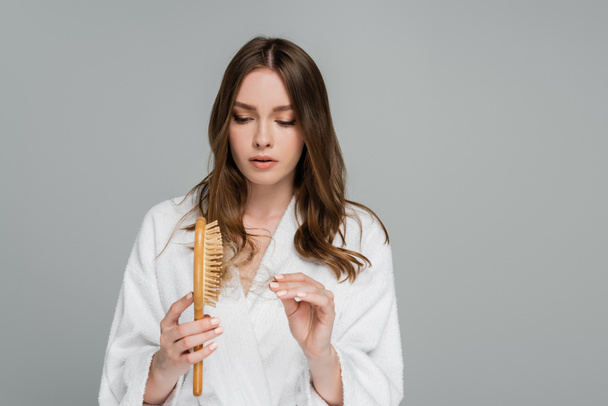 stressed young woman holding wooden hair brush and pulling damaged hair isolated on grey  - Photo, Image