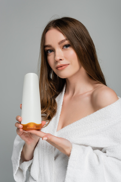 jonge vrouw met gezond haar holding fles met shampoo geïsoleerd op grijs - Foto, afbeelding
