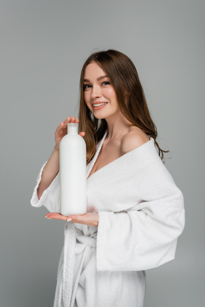 joyful young woman with shiny hair holding bottle with shampoo isolated on grey - Photo, Image