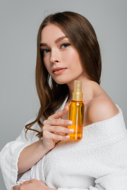 mujer joven con pelo brillante sosteniendo botella con aceite aislado en gris - Foto, Imagen