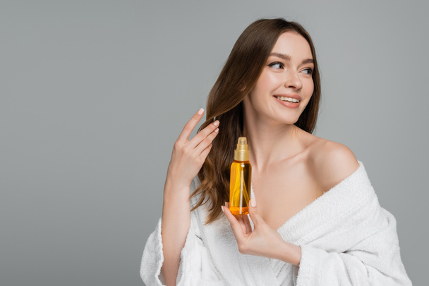 χαρούμενη νεαρή γυναίκα κρατώντας μπουκάλι με λάδι μαλλιών απομονώνονται σε γκρι - Φωτογραφία, εικόνα