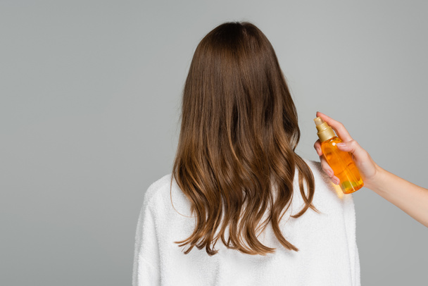 プロのヘアスタイリストは灰色に隔離された光沢のある髪を持つ若い女性の近くに油で瓶を保持 - 写真・画像