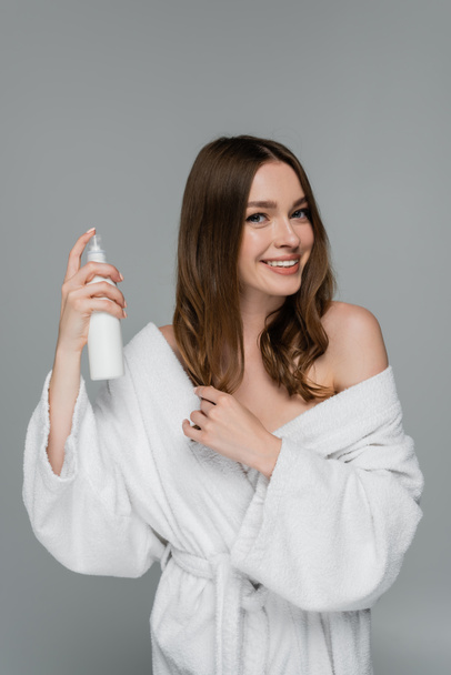 fröhliche junge Frau Styling glänzende Haare mit Haarspray isoliert auf grau  - Foto, Bild