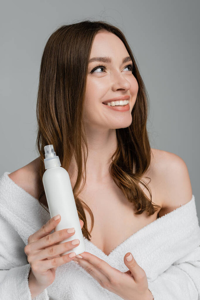 glimlachende jonge vrouw met glanzend haar houden haar spray geïsoleerd op grijs  - Foto, afbeelding