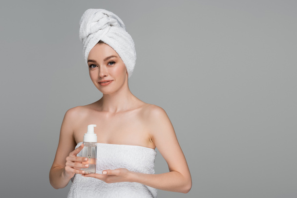 lächelnde junge Frau mit nackten Schultern und Handtuch auf dem Kopf, Flasche mit Reinigungsschaum isoliert auf grau  - Foto, Bild