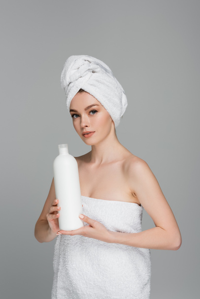 young woman with bare shoulders and towel on head holding bottle with hair conditioner isolated on grey  - Photo, Image