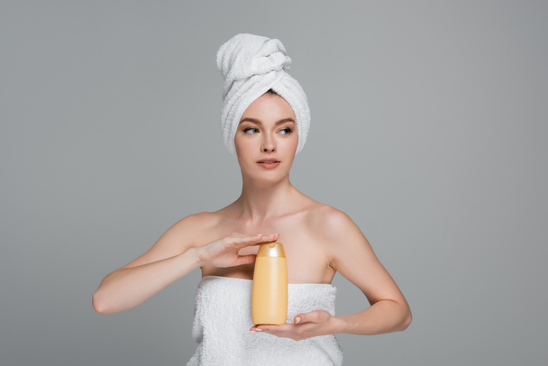 young woman with bare shoulders and towel on head holding bottle with shampoo isolated on grey  - Foto, Imagem