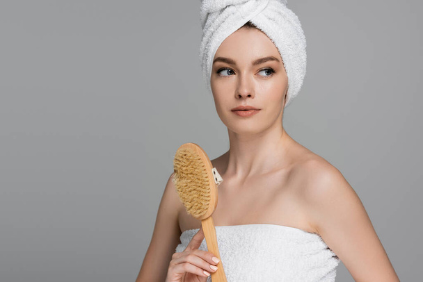 mujer joven con toalla en la cabeza sosteniendo cepillo de pelo de madera aislado en gris  - Foto, Imagen