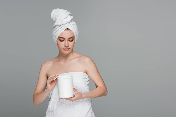 młoda kobieta z ręcznikiem na głowie trzymając pojemnik z maską do włosów izolowane na szaro  - Zdjęcie, obraz
