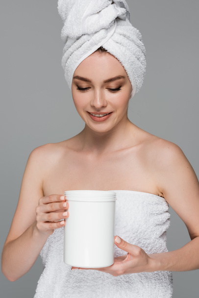 joyeuse jeune femme avec serviette sur la tête tenant conteneur avec masque capillaire isolé sur gris  - Photo, image
