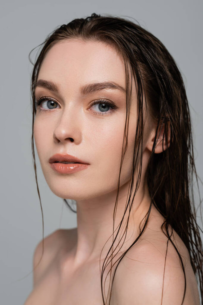 Porträt einer hübschen und jungen Frau mit nassen Haaren und nackten Schultern, die isoliert auf grau in die Kamera blickt  - Foto, Bild