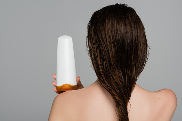 Rückansicht einer jungen Frau mit nassen Haaren und nackten Schultern, die eine Flasche mit Shampoo hält, isoliert auf grau  - Foto, Bild