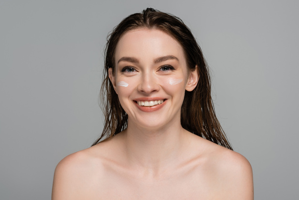 fröhliche junge Frau mit nassen Haaren und Feuchtigkeitscreme auf den Wangen vereinzelt auf grau  - Foto, Bild