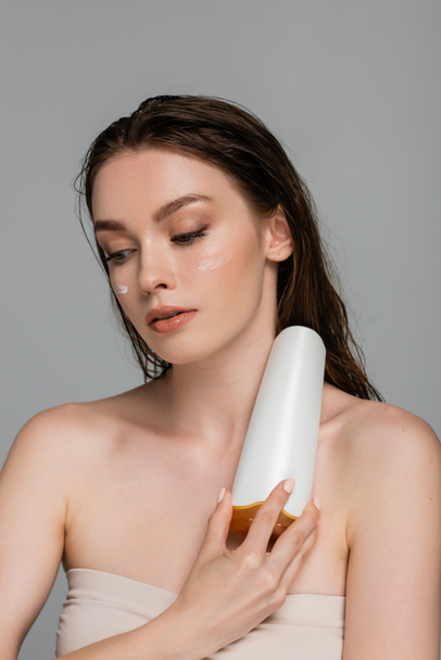 bonita mujer joven con el pelo mojado y crema hidratante en las mejillas sosteniendo botella con champú aislado en gris  - Foto, Imagen