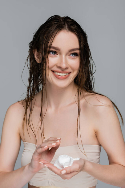 νέα και χαμογελαστή γυναίκα με βρεγμένα μαλλιά κρατώντας αφρό στο χέρι απομονώνονται σε γκρι - Φωτογραφία, εικόνα