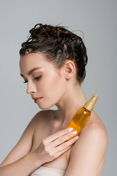 morena joven con cabello mojado sosteniendo botella con aceite de tratamiento aislado en gris  - Foto, Imagen