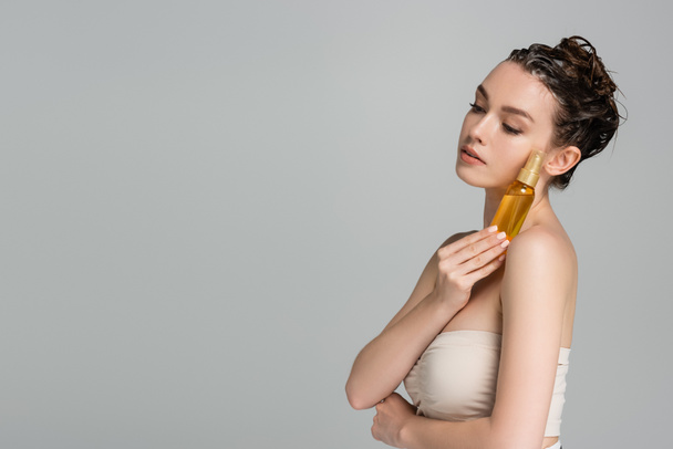 брюнетка молодая женщина с мокрыми волосами держа бутылку с маслом изолированы на сером  - Фото, изображение