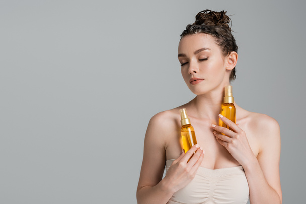 μελαχρινή νεαρή γυναίκα με κλειστά μάτια κρατώντας μπουκάλια με λάδι μαλλιών θεραπεία απομονώνονται σε γκρι  - Φωτογραφία, εικόνα
