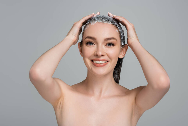 Ευτυχισμένη νεαρή γυναίκα με γυμνούς ώμους εφαρμογή μάσκα σε βρεγμένα μαλλιά απομονώνονται σε γκρι  - Φωτογραφία, εικόνα